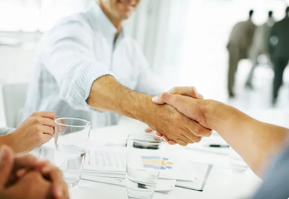 Close-up of a handshake between two people across a meeting table with documents and glasses of water, signifying a business agreement or successful negotiation